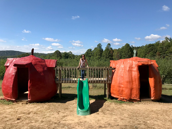 Playground at Sky Top Orchard in Flat Rock NC with brunette white woman standing on a wooden bridge between a red apple hut and orange pumpkin hut