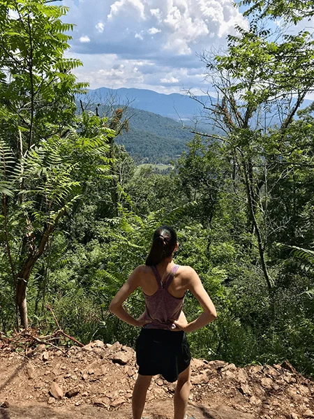 Craven Gap Hiking Trail with white brunette woman in workout skirt and top looking out over Blue Ridge Mountains with hands on hip