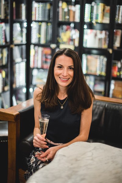 Battery Park Book Exchange Asheville Bookstore interior with bookshelves with clear glass and white brunette woman sitting on a couch wearing a blue dress and drinking a literary champagne cocktail