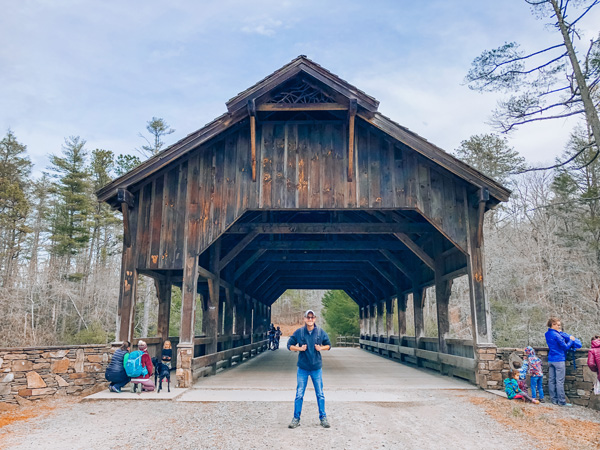 Asheville Waterfall Hikes Covered Bridge (wooden) at High Falls in DuPont Forest with white male hiking standing in front with hat and backpack