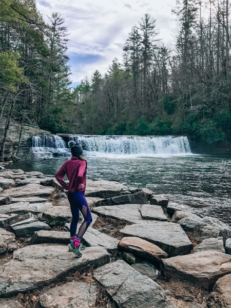 Hooker Falls at DuPont State Recreational Forest brunette white woman in pink and purple hiking gear in front of a waterfall near Asheville NC