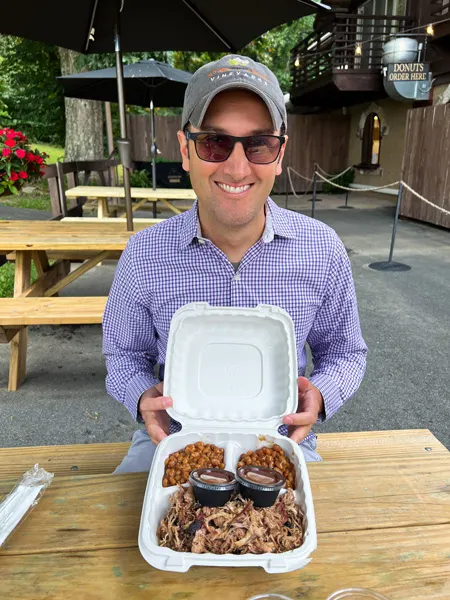 Jeter Mountain Farms 1813 Smokehouse Hendersonville with white male in sunglasses, hat, and purple long sleeved button down shirt with take away container of BBQ pulled pork at picnic table with two sides of baked beans