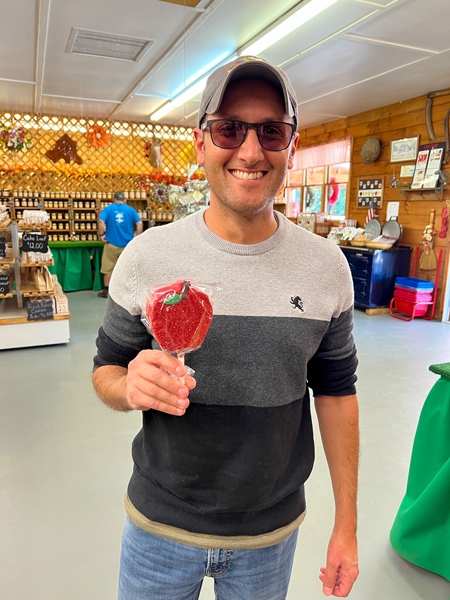 Mountain Fresh Orchard Hendersonville NC Store with white male in tan hat and ombre gray sweater holding an apple rice krispie treat