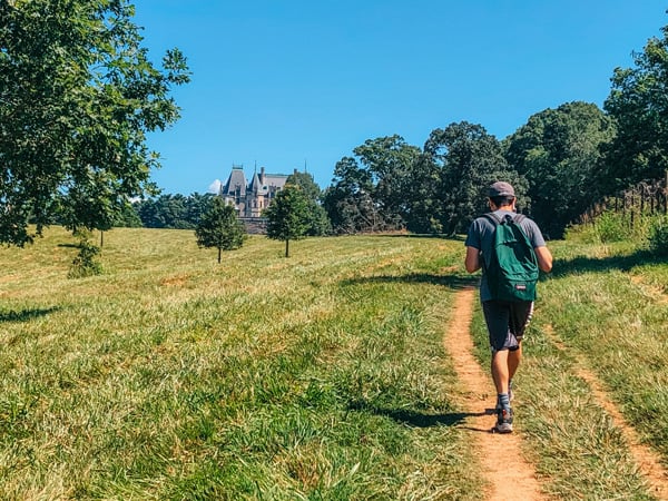Hiking in Asheville, NC with image of Biltmore Estate's Deerpark Trail with white brunette man hiking up to side of Biltmore with green backpack