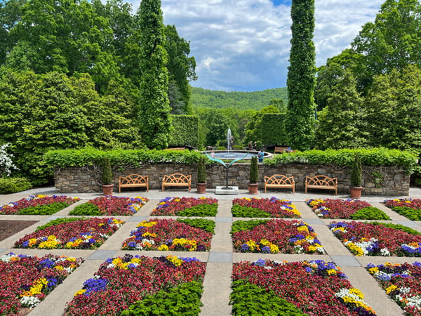 The NC Arboretum's Quilt Garden with rows of curated flowers in a quilt pattern with trees and fountain in background with blue cloudy sky