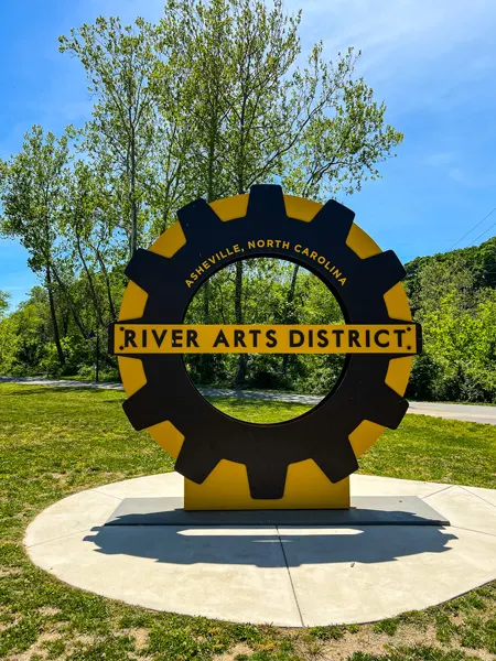 RAD Riverwalk Asheville NC with big yellow and black gear saying River Arts District Asheville, NC with walking path and green grass behind it
