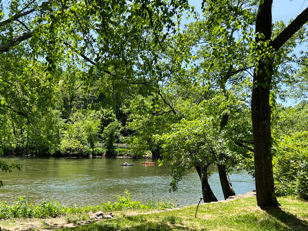 River Arts District Riverwalk French Broad River Asheville with river, green trees and grass, and people kayaking on the river