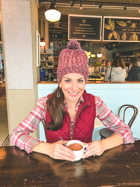 French Broad Chocolate Lounge in Asheville, NC with brunette white female wearing a red hat and vest with a cup of vegan hot chocolate