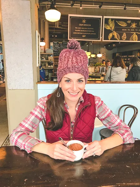 French Broad Chocolate Lounge in Asheville, NC with brunette white female wearing a red hat and vest with a cup of vegan hot chocolate