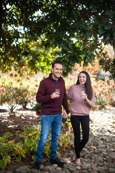 Burntshirt Vineyards Hendersonville NC with white brunette male and female drinking wine and walking. He is wearing a maroon sweater and she is in a pink top with black pants.