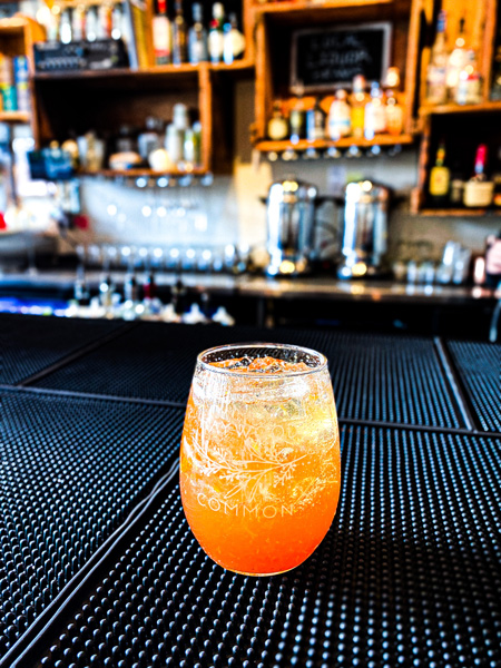 Haywood Common West Asheville Cocktail on bar with glasses and liquor in background. Cocktail is orange colored old fashioned with ice