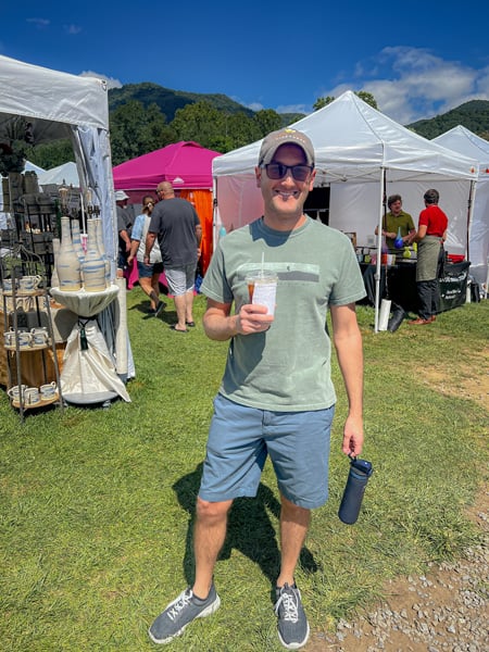 Maggie Valley Arts and Crafts Festival in September near Asheville with white brunette male wearing green shirt, blue shorts, hat, and sun glasses while carrying iced coffee in front of outdoor vendors