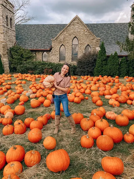 Pumpkin Patch with white female in jeans and pink top holding a white pumpkin with church in the background