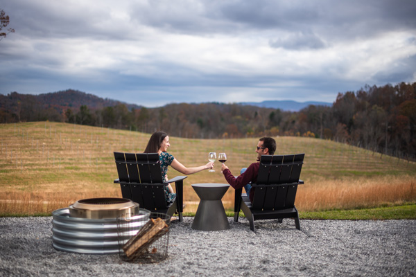 Wineries In and Near Asheville Marked Tree Vineyard with brunette white male and female sitting in chairs looking out at the mountains doing a cheers with glasses of wine