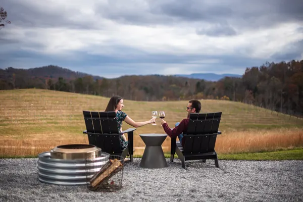 Wineries In and Near Asheville Marked Tree Vineyard with brunette white male and female sitting in chairs looking out at the mountains doing a cheers with glasses of wine