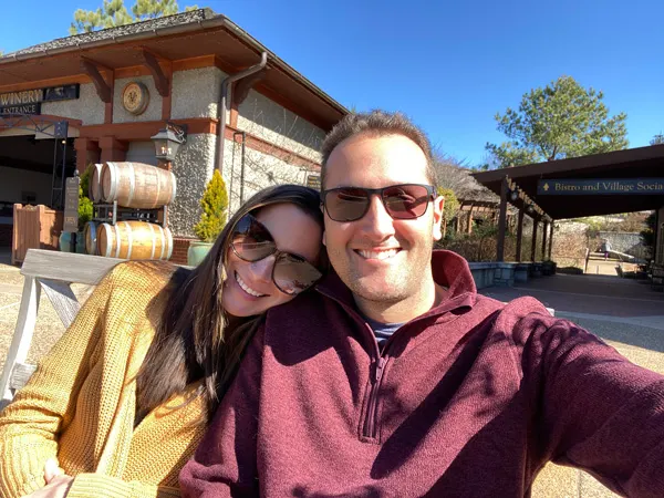 Antler Hill Village Biltmore Estate with white brunette male and woman taking a selfie in front of winery on a sunny day; he's wearing a maroon sweater and she's in a mustard yellow sweater