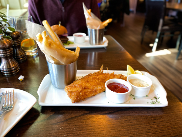 Cedrics Tavern Restaurant Biltmore Estate lunch with fish and chips on long white plate with fries, tarter sauce, and ketchup on table with blurred male wearing purple in the background