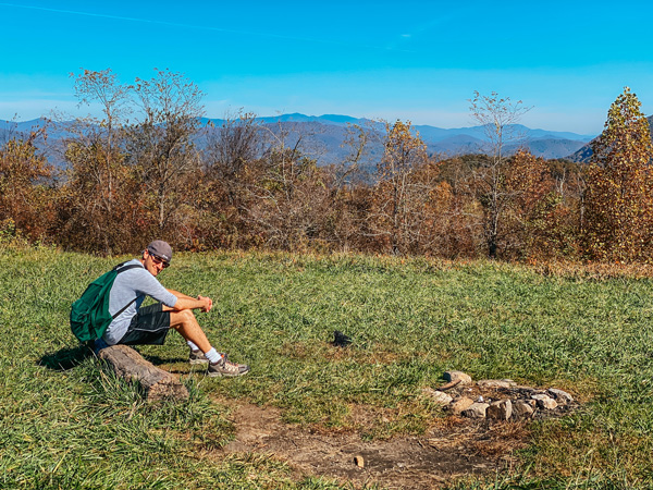 Picnic Spot At Trombatore Trail Blue Ridge Pastures with brunette white male wearing a green hiking backpack sitting on a logPicnic Spot At Trombatore Trail Blue Ridge Pastures