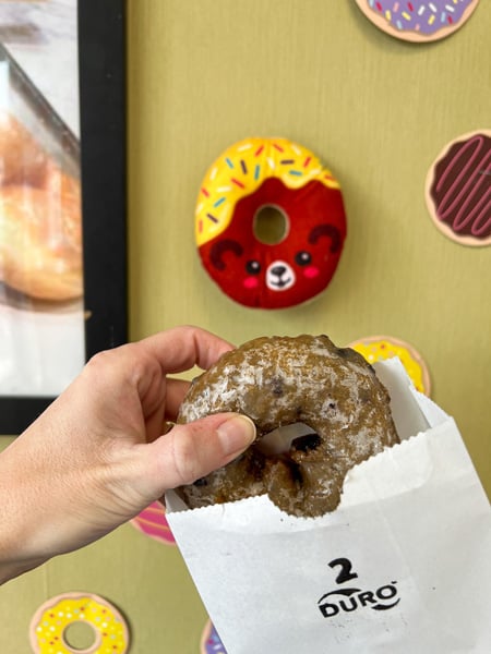 Avas Donuts In Asheville North Carolina with white hand holding up glazed blueberry donut in front of wall decorated with fake donuts with faces