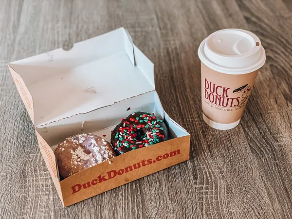 Duck Donuts Asheville to go box with two sprinkled and glazed donuts in it with to-go cup of coffee on light gray brownish table