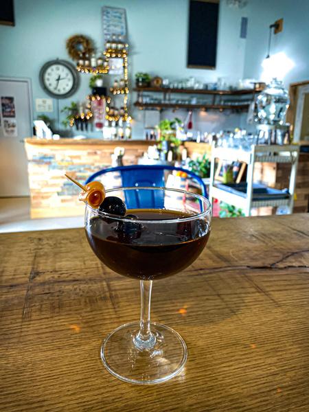 Oak And Grist Distilling Company Black Mountain NC with red-brown cocktail on wooden table with bar and clock in background