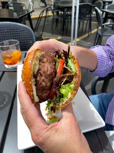 Rye Knot Burger Asheville NC with white hands holding huge beef burger with lettuce and tomato on gluten free bun and Old Fashioned cocktail in background