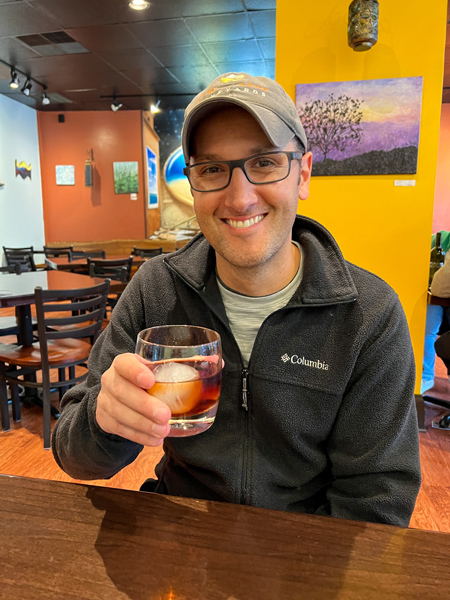 Blue Dream Curry House Asheville brown cocktail with ball ice cube in hand of white male in hat and black fleece with paintings on wall in background