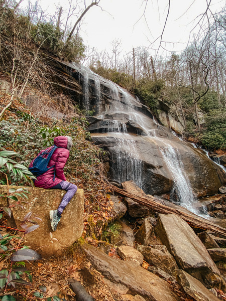 Daniel Ridge Falls Winter Hike Pisgah National Forest with woman in plum colored coat with backpack sitting in front of Daniel Ridge Falls