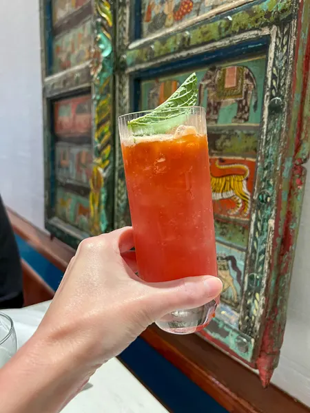 Mehfil Indian Restaurant Asheville NC red cocktail being held up by white hand in front of colorful wall decor and cocktail has a basil garnish