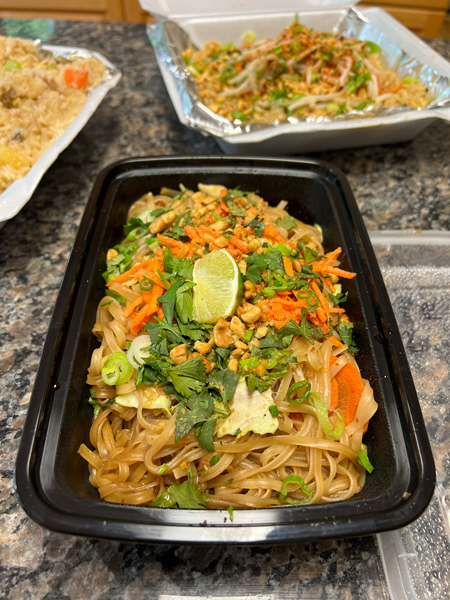 Thai Pearl Takeout Asheville NC with gluten free pad Thai noodles with carrots, lime, and cilantro in black takeout container on marble kitchen island with blurred takeout dishes with noodles in background