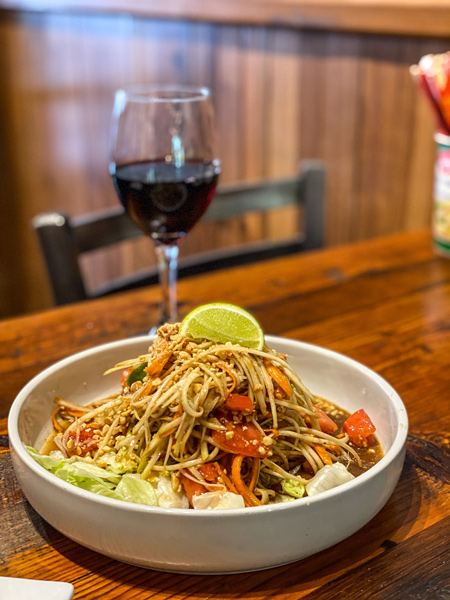 Thai Pearl Thai Papaya Salad with salad in white bowl garnished with lime and glass of wine in the background on a brown table