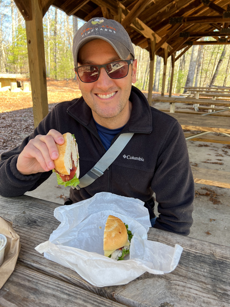 Triple Falls Picnic Area at DuPont State Forest in NC with white brunette make wearing hat, fleece, and sunglasses eating a takeout sub at covered picnic table