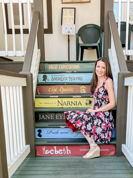White brunette woman with long hair in pink and black flowering dress sitting on stairs labeled with book spines at The Book And Bee Cafe and Tea in Hendersonville NC