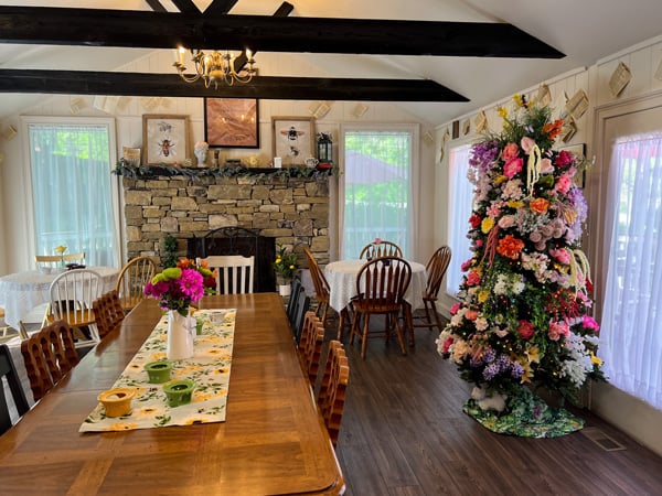 The Book and Bee Cafe Hendersonville North Carolina with long family-style table in a large white room with wood beam ceilings, a stone fire place, and a tree decorated with spring colors like pinks and lavender
