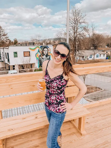 Archetype Brewing West Asheville with white brunette female wearing flowered shirt, jeans, and sunglasses holding a white ale in front of street art on a rooftop seating area