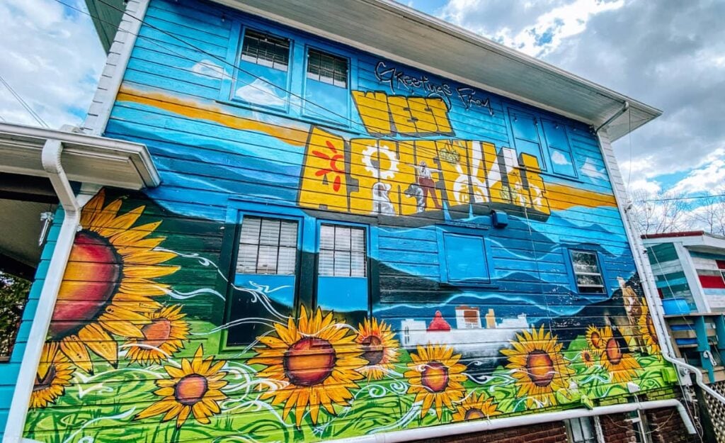 Featured image for the article, "Best Neighborhoods in Asheville, NC" with photograph of mural on the side of a house that says "West Asheville" in yellow with blue sky and yellow and orange sunflowers in green grass