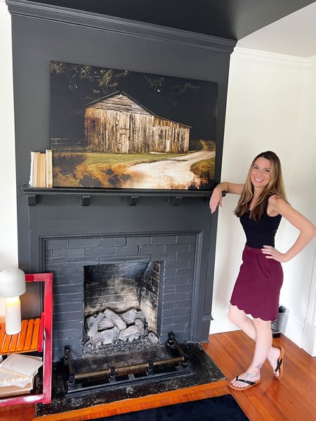 Gray fireplace with Christine, a white brunette woman in red skirt and black top standing in front of it, at the Blind Tiger in Asheville,North Carolina