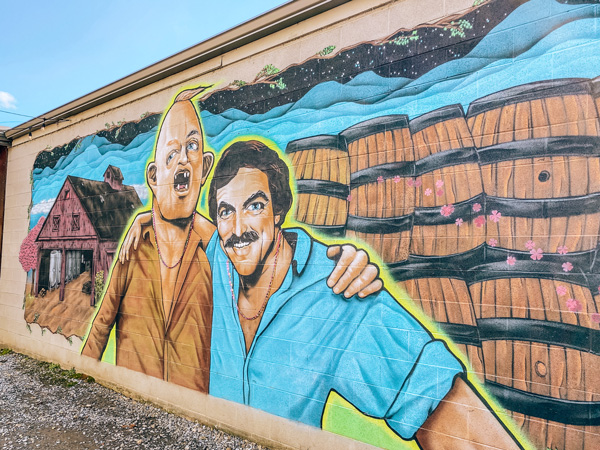 Burial Beer Co Tom Selleck Sloth Mural Asheville with Sloth's arm around Selleck next to barrels and a barn