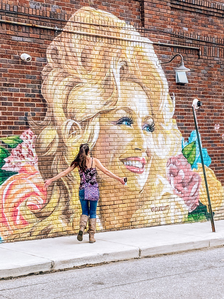 Dolly Parton Mural West Asheville Neighborhood with white brunette woman running up to Dolly Parton face on a street mural