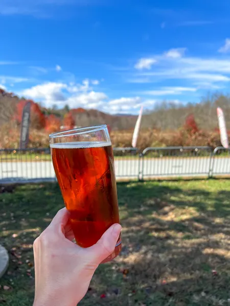 Oskar Blues Brewery Brevard NC with white hand holding amber-orange Oktoberfest beer at picnic table with fall foliage in the background and blue sky