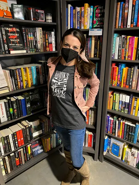 Sassafras On Sutton Bookstore Black Mountain with white brunette female in pink jacket and jeans wearing a mask standing among black bookshelves filled with books