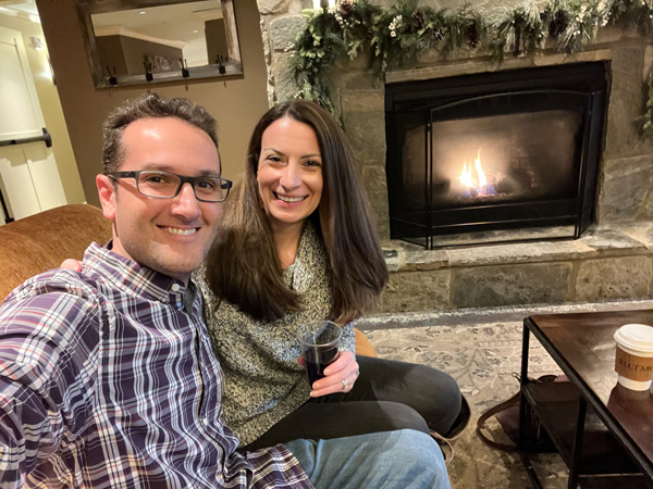 Christine and Tom, a white brunette male and female, on couch taking a selfie in front of lit gas fireplace at the Village Hotel on Biltmore Estate in Asheville, NC