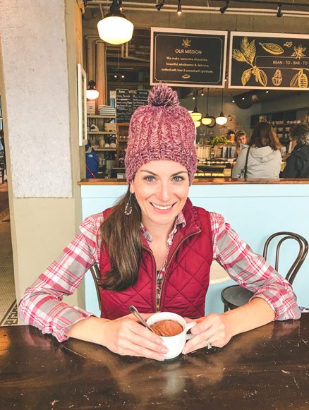 Best Things To Do In Asheville French Broad Chocolate Lounge with brunette white female wearing a red hat and vest with a cup of vegan hot chocolate