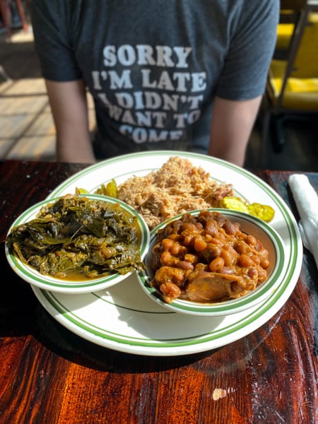 Buxton Hall BBQ Asheville NC with white male burred in background in blue t-shirt with plate of collard greens, baked beans, and BBQ pulled pork in front of him