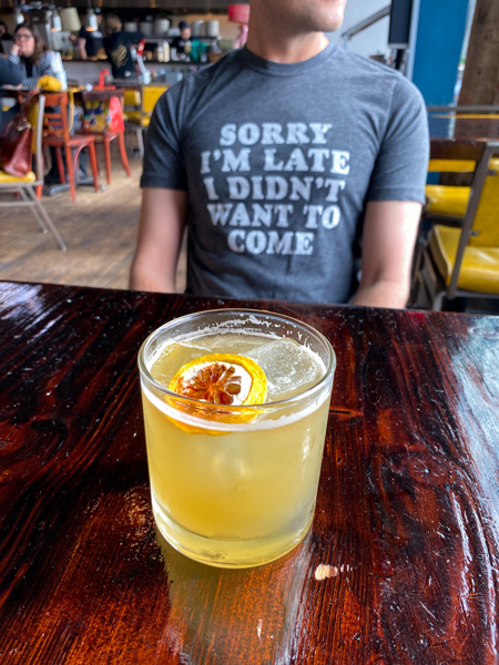 Buxton Hall Barbecue Downtown Asheville NC with man blurred in the background wearing blue shirt with yellow cocktail with lemon garnish sitting on dark brown table