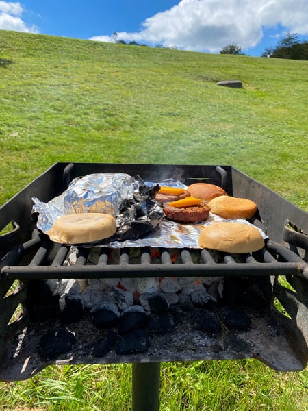 Craggy Gardens Picnic Area Charcoal Grill with burgers and buns on foil on grill with green grass and blue sky