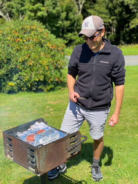 Tom, a white brunette male in black fleece and cargo shorts, grilling burgers on charcoal grill at Craggy Gardens for a picnic