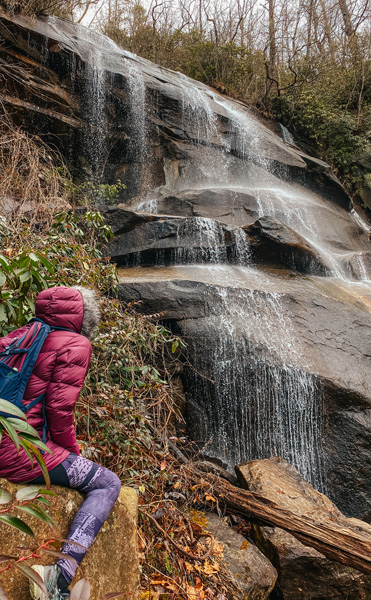 Daniel Ridge Falls Trail with woman in garnet colored winter coat with hood and purple hiking leggings sitting on a rock and looking at waterfall