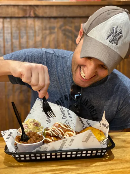 Moe's Original BBQ with white brunette male wearing a NY Yankees hat eating smoked chicken from a basket with sides like baked beans and potato salad