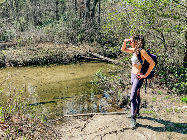 Pink Beds Hike Pisgah Forest with white brunette woman in purple leggings, sneakers, and sports bra wearing a backpack looking out at stream